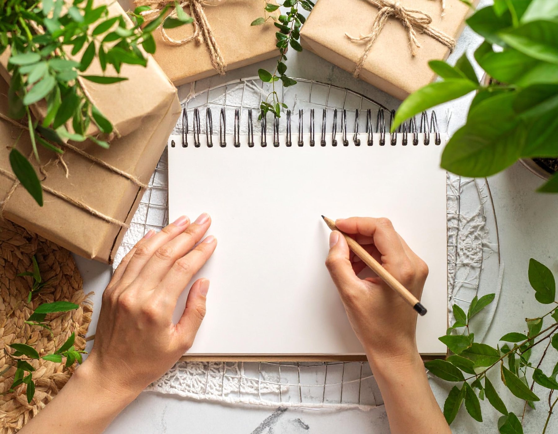 A spiral-bound sketchbook open on a desk beside plants and a pencil